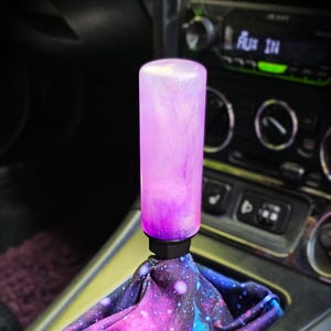 Op de afbeelding: Een cilindrische versnellingspookknop met een doorschijnende roze en paarse glitterafwerking, bevestigd aan een zwarte basis. De versnellingspookhoes heeft een levendige sterrenstelselprint met tinten diepblauw, magenta en witte sterachtige spikkels.