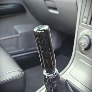 May include: A cylindrical gear shift knob with a glossy black finish and subtle silver glitter flecks. The knob features a chrome-coloured metal base collar and is attached to a black leather-textured shift boot inside a vehicle interior.
