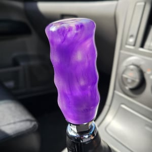 May include: A custom purple shift knob with a swirling, marbled effect. The knob is mounted on a black leather shift boot and features a chrome base. The interior of a car is visible in the background.