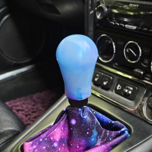 May include: A car gear shift knob featuring a smooth, light blue, bulbous handle attached to a black base, paired with a fabric shift boot printed with a vibrant purple, pink, and blue galaxy pattern featuring scattered white stars.