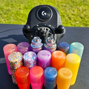 Pode incluir: Uma coleção de botões de câmbio coloridos e brilhantes para um carro. Os botões estão dispostos em semicírculo em torno de um volante de jogo preto com um logotipo G.