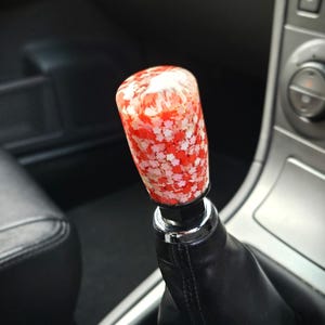 Peut inclure: Un pommeau de levier de vitesse cylindrique doté d'un corps en résine transparente incrusté de nombreuses petites confettis en forme d'étoile dans des tons de rouge, rose et blanc. Il est fixé à un soufflet en cuir noir avec une base argentée.