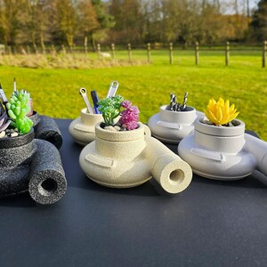 May include: Three decorative turbocharger-shaped planters, each in a different colour: black, white and beige. Each planter has a small succulent plant inside. The black planter has two pens and a pencil inside. The beige planter has three pens inside. The white planter has a few small drill bits inside.