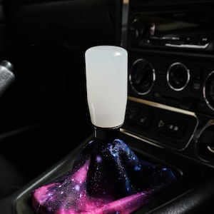 May include: A cylindrical white gear shift knob attached to a black metal base, featuring a fabric shift boot decorated with a vibrant galaxy print in shades of deep purple, blue, and bright pink, installed in a vehicle center console.