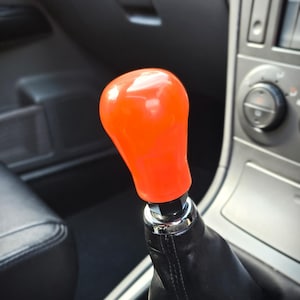 Peut inclure: Un pommeau de levier de vitesse orange vif, lisse et bulbeux, fixé à un soufflet en cuir noir. Le pommeau présente une finition brillante et est monté sur une base métallique, situé dans la console centrale d'un habitacle de véhicule.