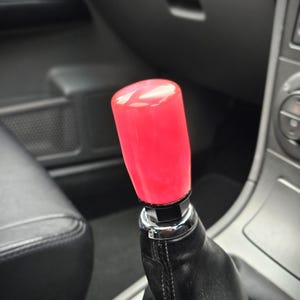 Könnte beinhalten: Ein leuchtend roter Schaltknauf mit zylindrischer Form. Der Knauf ist auf einem schwarzen Lederschaltsack montiert, mit einem Chromzierring. Der Innenraum eines Autos ist im Hintergrund zu sehen.