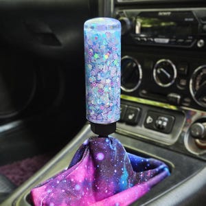 May include: A cylindrical gear shift knob filled with light blue liquid, iridescent glitter, and small flower-shaped sequins, attached to a black base. The gear shift boot features a vibrant galaxy print with shades of purple, pink, and blue.