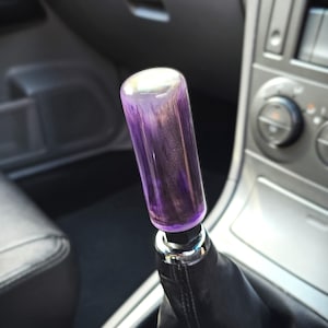 May include: A cylindrical gear shift knob featuring a translucent purple and black marbled resin finish. The knob is mounted on a black leather shift boot with a chrome-coloured base ring, positioned within the centre console of a vehicle.