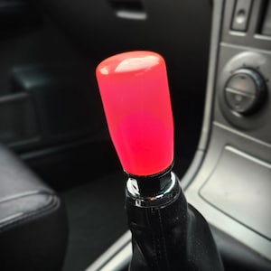 May include: A bright red shift knob with a cylindrical shape, mounted on a black leather shift boot. The knob is illuminated, and the interior of a car is visible in the background.