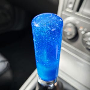 Pode incluir: Manípulo de mudança de velocidades de carro personalizado, com um acabamento em resina azul vibrante com glitter. O botão cilíndrico tem uma aparência polida e brilhante, refletindo a luz e adicionando um toque de brilho ao interior do veículo.