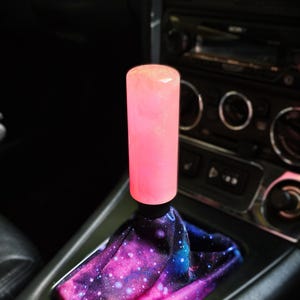 May include: A cylindrical gear shift knob with a translucent, light pink, glittery finish attached to a black base. The gear shifter is covered by a fabric boot featuring a vibrant galaxy print with shades of purple, blue, and pink with white stars.