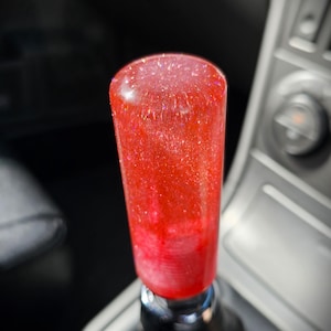 Peut inclure: Un pommeau de levier de vitesses rouge infusé de paillettes. Le bouton cylindrique présente un effet de dégradé, passant d'un sommet plus clair et rempli de paillettes à un rouge plus profond à la base. Le bouton est monté sur un levier chromé, sur un intérieur flou.