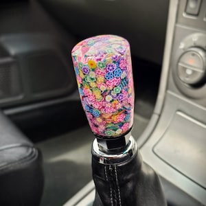 Könnte beinhalten: Ein dekorativer Schalthebelknauf mit einem bunten Blumenmuster. Der zylindrische Knauf zeigt rosa, blaue, gelbe und grüne Blumen. Er ist auf einem schwarzen Lederschaltsack montiert. Der Hintergrund zeigt das Autointerieur.