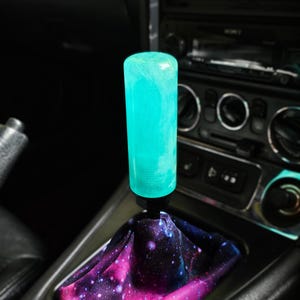 May include: A cylindrical gear shift knob with a translucent, glowing turquoise finish, mounted on a black base. The gear shift boot features a vibrant galaxy print with deep purple, pink, and blue nebula patterns and scattered white star details.