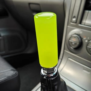 Può includere: Un pomello del cambio verde neon brillante. La manopola cilindrica è montata su una base cromata e un soffietto del cambio in pelle nera. L'interno di un'auto è visibile sullo sfondo.