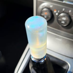 May include: A pearlescent, iridescent shift knob with a cylindrical design. The knob has a clear, shimmering appearance with hints of blue and yellow. It is mounted on a black leather shift boot in a vehicle's interior.