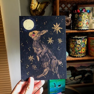 May include: A black card with a gold rabbit looking up at a full moon. The background is a dark blue with gold stars.