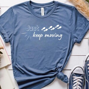 Peut inclure: T-shirt bleu avec l'inscription blanche &laquo;&nbsp;Just keep moving&nbsp;&raquo; et des empreintes de pas blanches. Le t-shirt est nou&eacute; &agrave; la taille. Des baskets bleues sont visibles.