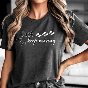 May include: Dark gray t-shirt with the text "Just keep moving" in white script. The shirt features a minimalist design with footprints above the words. The short-sleeved tee is made of a soft material.