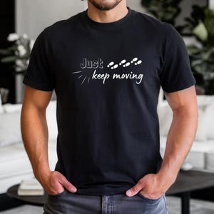 Peut inclure: T-shirt noir avec l'inscription blanche "Just keep moving" et un motif de pas. Le t-shirt &agrave; manches courtes semble &ecirc;tre en coton. Le design est centr&eacute; sur la poitrine.