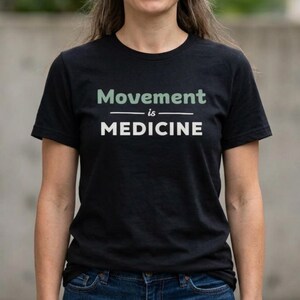 May include: Black t-shirt with the text "Movement is Medicine" in white and green lettering. The shirt is a crew neck style and appears to be made of a soft material. The text is centered on the front of the shirt.
