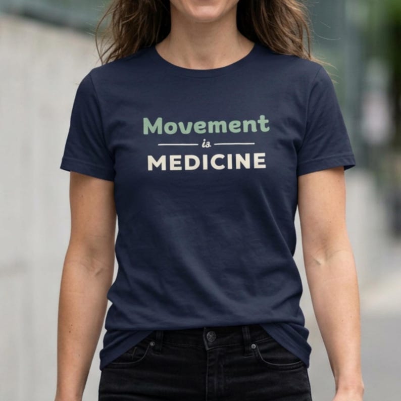 May include: Navy blue t-shirt with the text "Movement is Medicine" in white and light green. The shirt has short sleeves and a crew neck. The text is centered on the front of the shirt.
