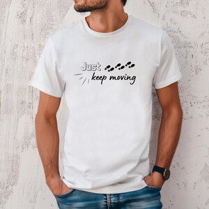 Peut inclure: T-shirt blanc avec l'inscription "Just keep moving" en noir, accompagn&eacute;e d'empreintes de pas noires. Le t-shirt est &agrave; manches courtes et a un col rond.
