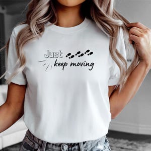Peut inclure: T-shirt blanc avec l'inscription noire &laquo; Just keep moving &raquo; et des empreintes de pas noires. Le t-shirt a des manches courtes et un col rond.