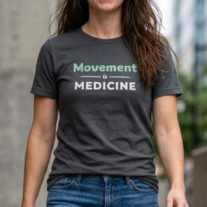 May include: Dark gray t-shirt with the text "Movement is Medicine" in white and green. The shirt is a crew neck style and is paired with blue jeans. The image is taken outdoors.