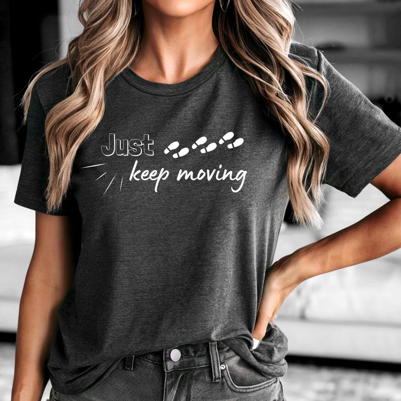 May include: Dark gray t-shirt with the text "Just keep moving" in white script. The shirt features a minimalist design with footprints above the words. The short-sleeved tee is made of a soft material.