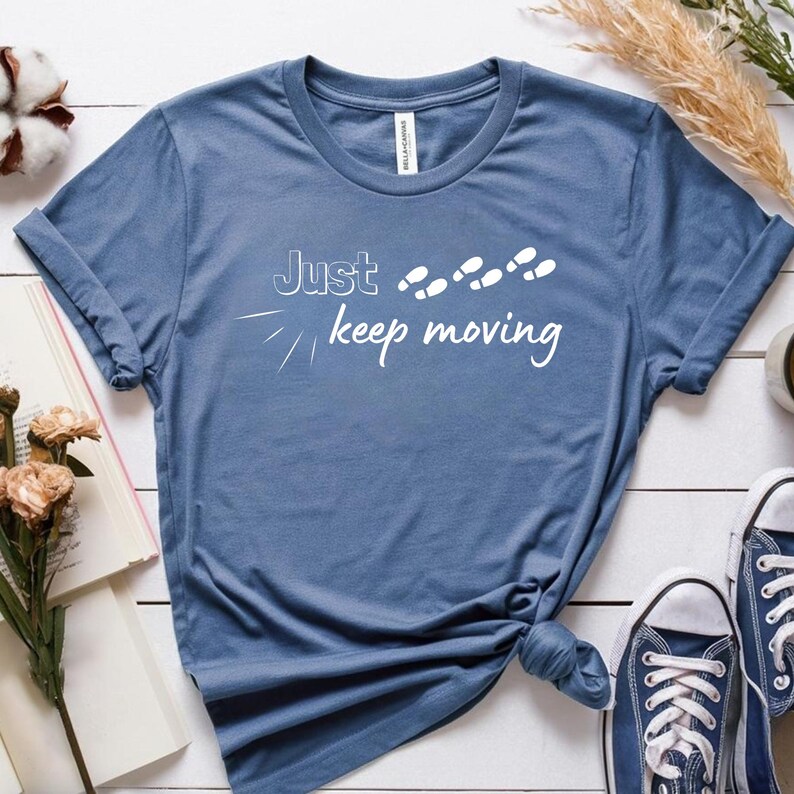 Peut inclure: T-shirt bleu avec l'inscription blanche &laquo;&nbsp;Just keep moving&nbsp;&raquo; et des empreintes de pas blanches. Le t-shirt est nou&eacute; &agrave; la taille. Des baskets bleues sont visibles.