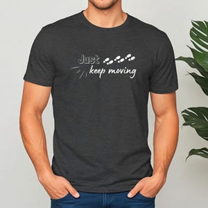 Peut inclure: T-shirt gris fonc&eacute; avec l'inscription blanche "Just keep moving". Le design comprend des empreintes de pas et des lignes stylis&eacute;es. Le t-shirt est &agrave; col rond et semble &ecirc;tre en mati&egrave;re douce.