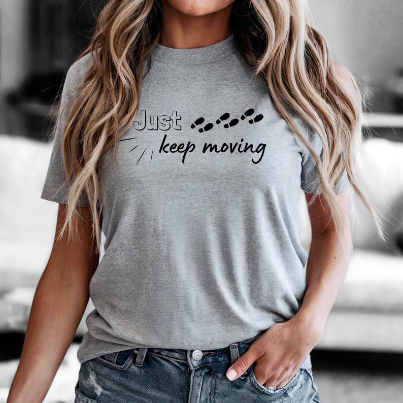 Peut inclure: Un t-shirt gris avec l'inscription "Just keep moving" en &eacute;criture noire et de petites empreintes de pas. Le t-shirt est de style col rond et est fait d'une mati&egrave;re douce.