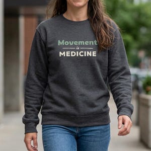 May include: Dark gray sweatshirt with the text "Movement is Medicine" in light green and white. The sweatshirt is a crew neck style and is paired with blue jeans. The overall theme is health and wellness.