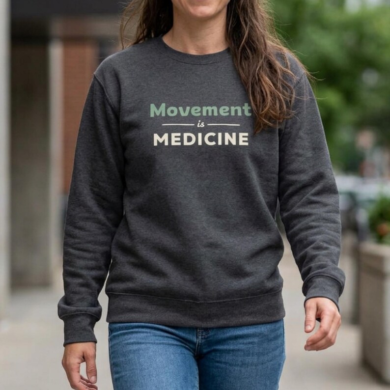 May include: Dark gray sweatshirt with the text "Movement is Medicine" in light green and white. The sweatshirt is a crew neck style and is paired with blue jeans. The overall theme is health and wellness.