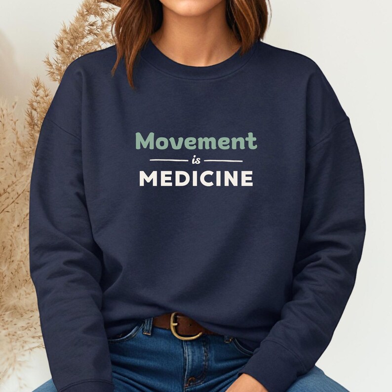 May include: Navy blue sweatshirt with the text "Movement is Medicine" in white and green. The sweatshirt has a crew neck and long sleeves. The model is wearing blue jeans and a brown belt.