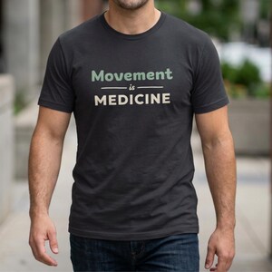 May include: Dark gray t-shirt with the text "Movement is Medicine" in green and white. The shirt has a crew neck and short sleeves. The shirt is made of a soft material.