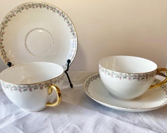 May include: Two teacups and saucers with a floral pattern and gold trim. The cups and saucers are white with a delicate pink and green floral design around the rim. The cups have gold handles.