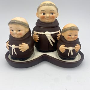 May include: A ceramic salt and pepper set featuring three monk figurines in brown robes. The figurines have smiling faces and are arranged on a brown and white tray. The set is designed for tabletop use.