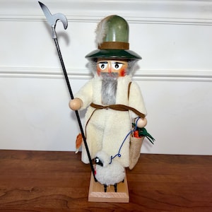 May include: A wooden nutcracker figure wearing a green hat with a feather, a long white coat, and a brown belt. The figure holds a tall black staff with a silver metal hook and carries a small burlap bag. A white sheep on a blue leash stands nearby.