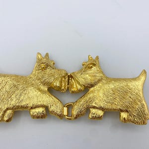 May include: Gold-toned metal belt buckle featuring two Scottie dogs facing each other, with their noses touching. The dogs are detailed with textured fur and have a realistic appearance.