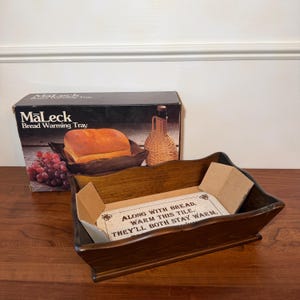 May include: A wooden bread warming tray with a dark finish. The tray has a rectangular shape with angled sides. A box with the text "MaLeck Bread Warming Tray" is behind the tray. The box shows a loaf of bread and grapes.