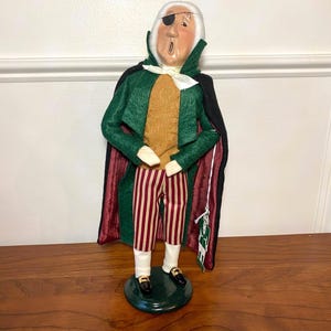 May include: A decorative figurine of a man with white hair, an eye patch, and a green coat with a black cape. He wears a gold vest, striped pants, and black shoes. The figurine stands on a green base.