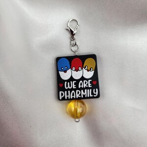 May include: A black charm with three cartoon pills, each with a different color face. The charm has a silver clip and a yellow bead. The text on the charm reads "We are Pharmily".