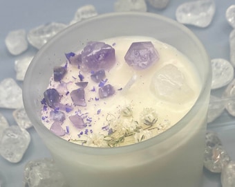 May include: A white candle in a frosted glass jar, decorated with small purple amethyst crystals and white dried flowers. The candle is surrounded by clear quartz crystals.