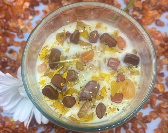 Puede incluir: Un tarro de vela de vidrio transparente con una vela blanca y cristales incrustados. La vela está rodeada de purpurina dorada y pequeños trozos de cristales amarillos y naranjas. El tarro está sentado sobre una superficie blanca con una dispersión de pequeñas piedras naranjas y rojas.