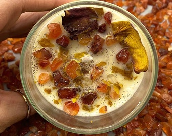Puede incluir: Un tarro de vela de vidrio transparente con una vela blanca. La vela está decorada con pequeñas piedras rojas y naranjas y una flor seca. El tarro está rodeado de pequeñas piedras rojas y naranjas.