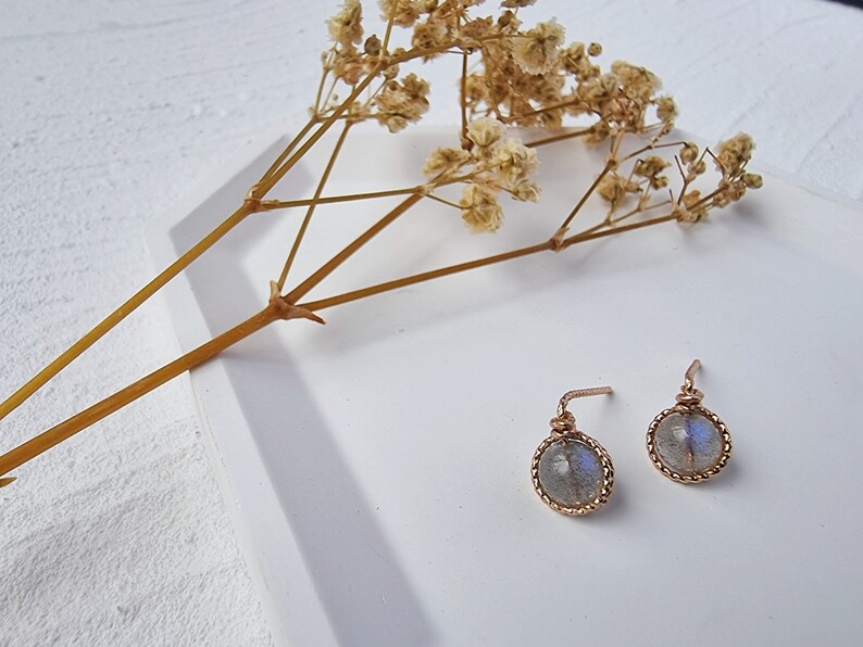 May include: A pair of silver earrings with a round, iridescent labradorite stone set in a gold-toned bezel. The earrings have a delicate, wire-wrapped design.