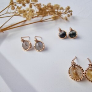 May include: Three pairs of gold-toned earrings with different gemstone accents. The first pair features a light gray stone, the second pair features a black stone, and the third pair features a yellow stone.