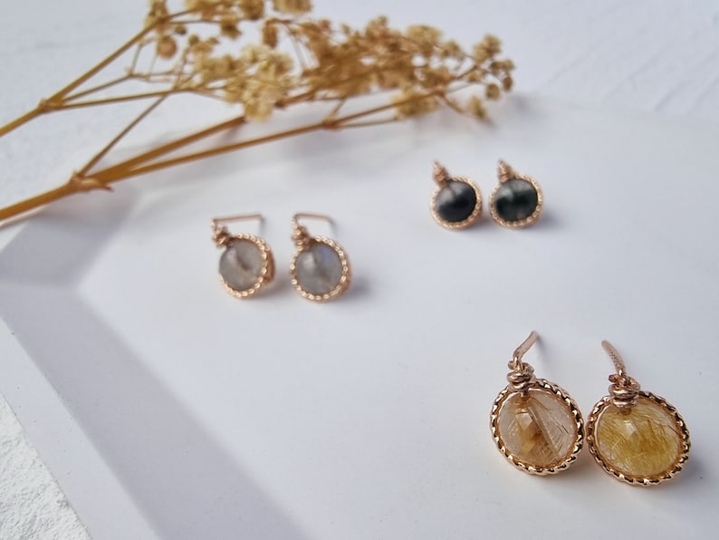 May include: Three pairs of gold-toned earrings with different gemstone accents. The first pair features a light gray stone, the second pair features a black stone, and the third pair features a yellow stone.